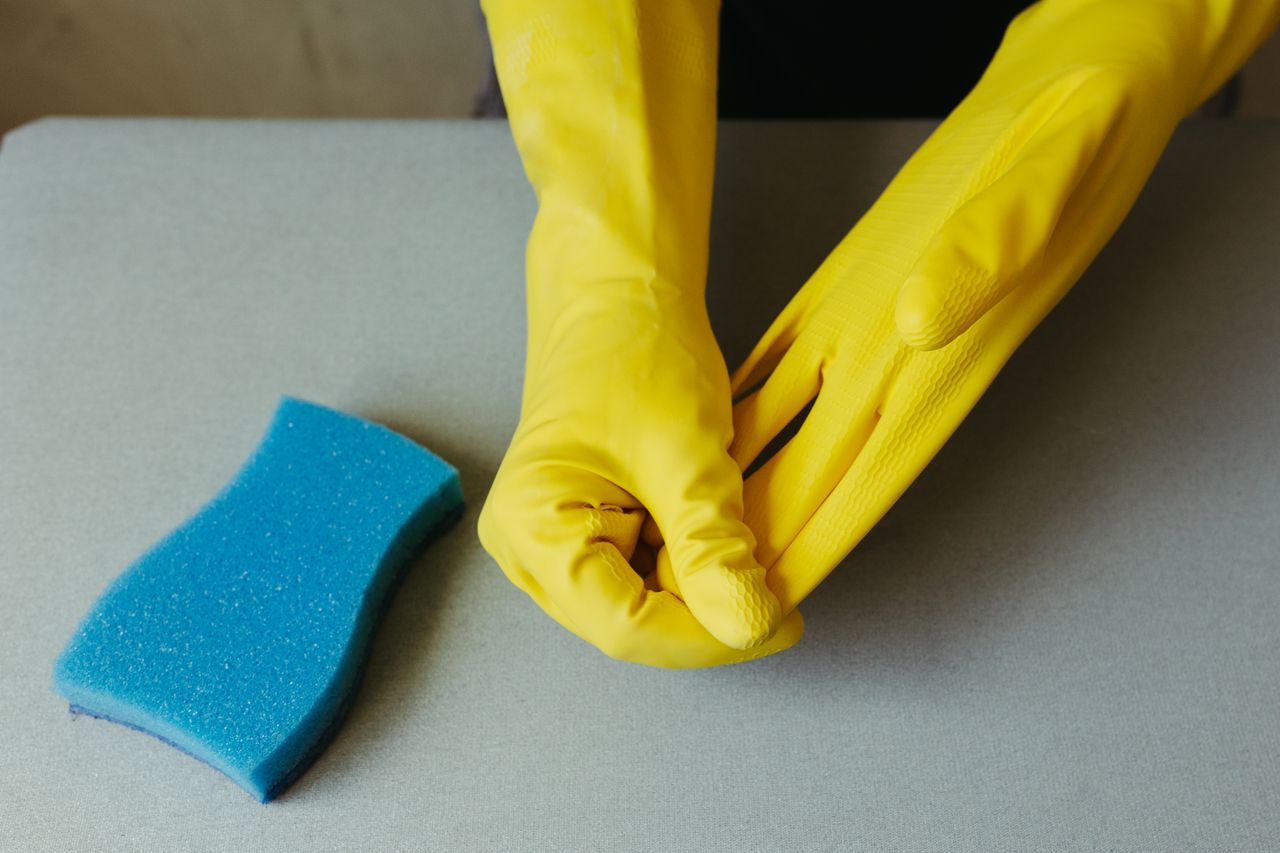 why-choose-us Close-up of hands wearing yellow gloves next to blue sponge on gray surface.